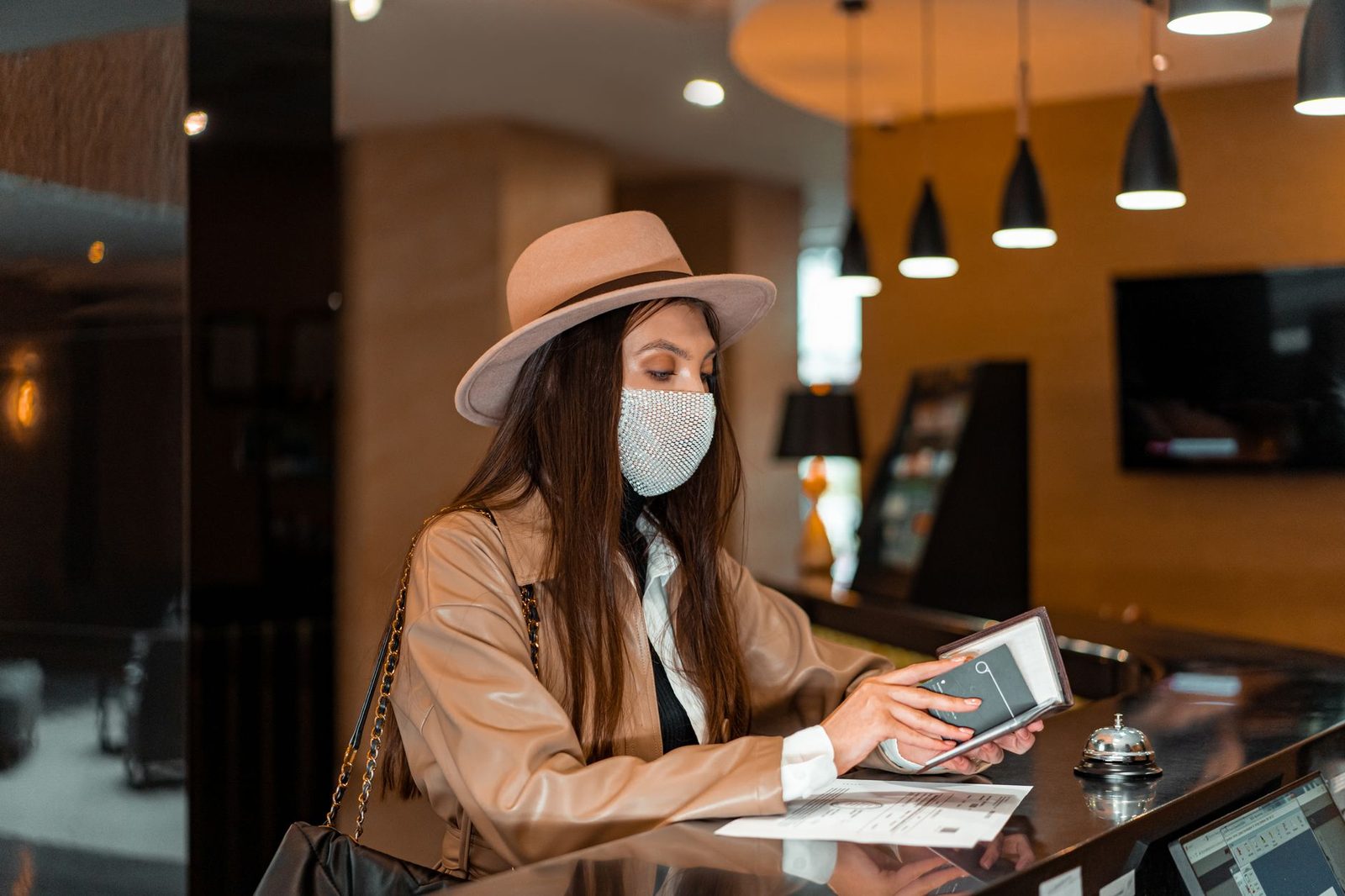 Woman wearing mask and hat checking in at hotel front desk, symbolizing travel and safety.