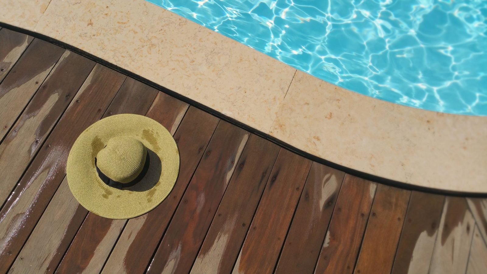 A straw hat rests on a wooden deck beside a sparkling blue swimming pool, perfect for summer relaxation.